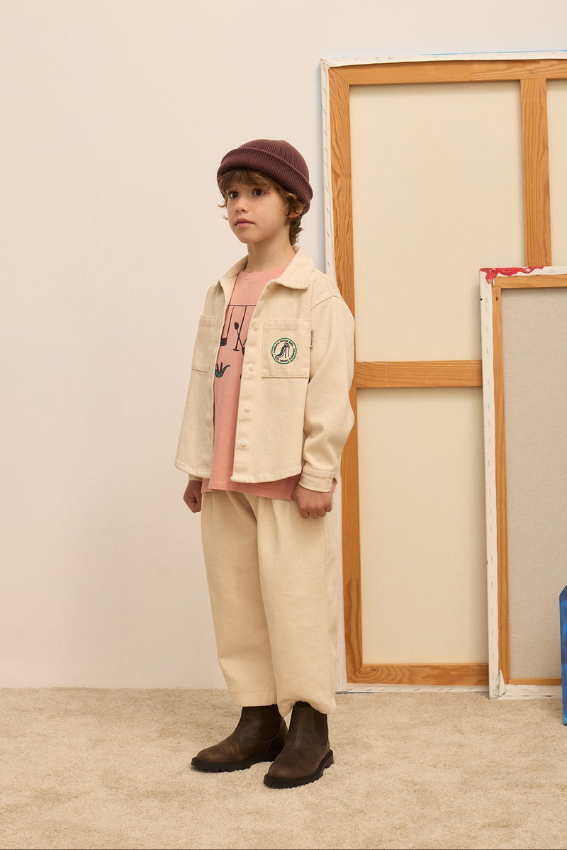 A child model wearing a beige denim jacket and matching trousers over a pink shirt, paired with a dark maroon beret and brown boots, standing in a minimalist studio setting.